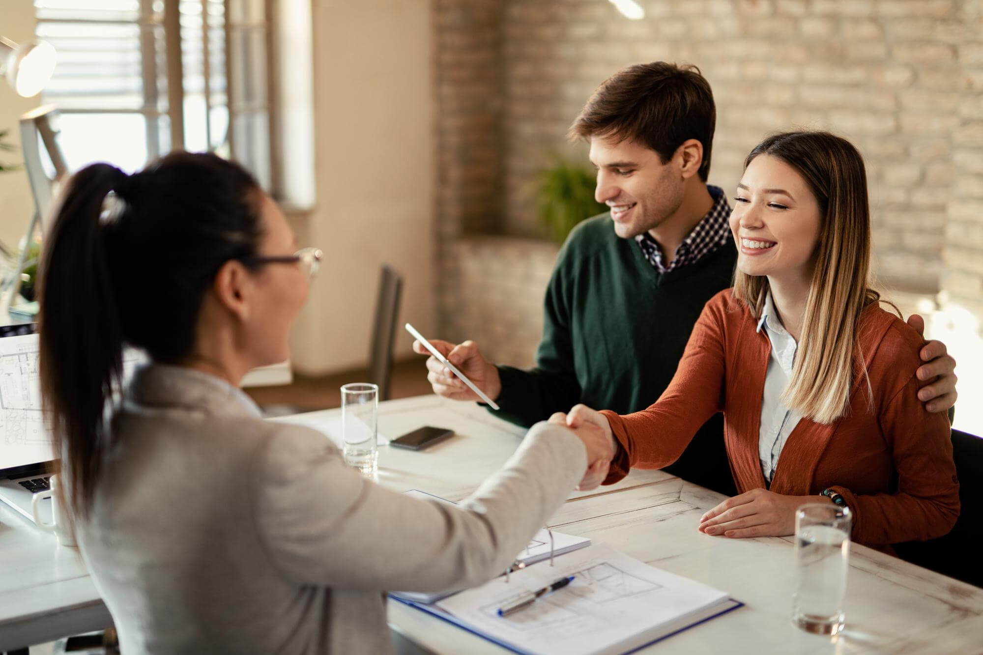Young happy woman shaking hands with real estate agent while her husband Young happy woman shaking hands with real estate agent while her husband