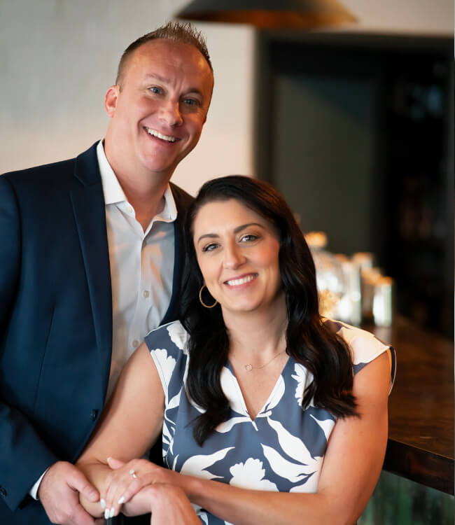 Smiling couple posing together indoors, with the man in a navy suit and the woman in a floral dress, leaning on a wooden counter